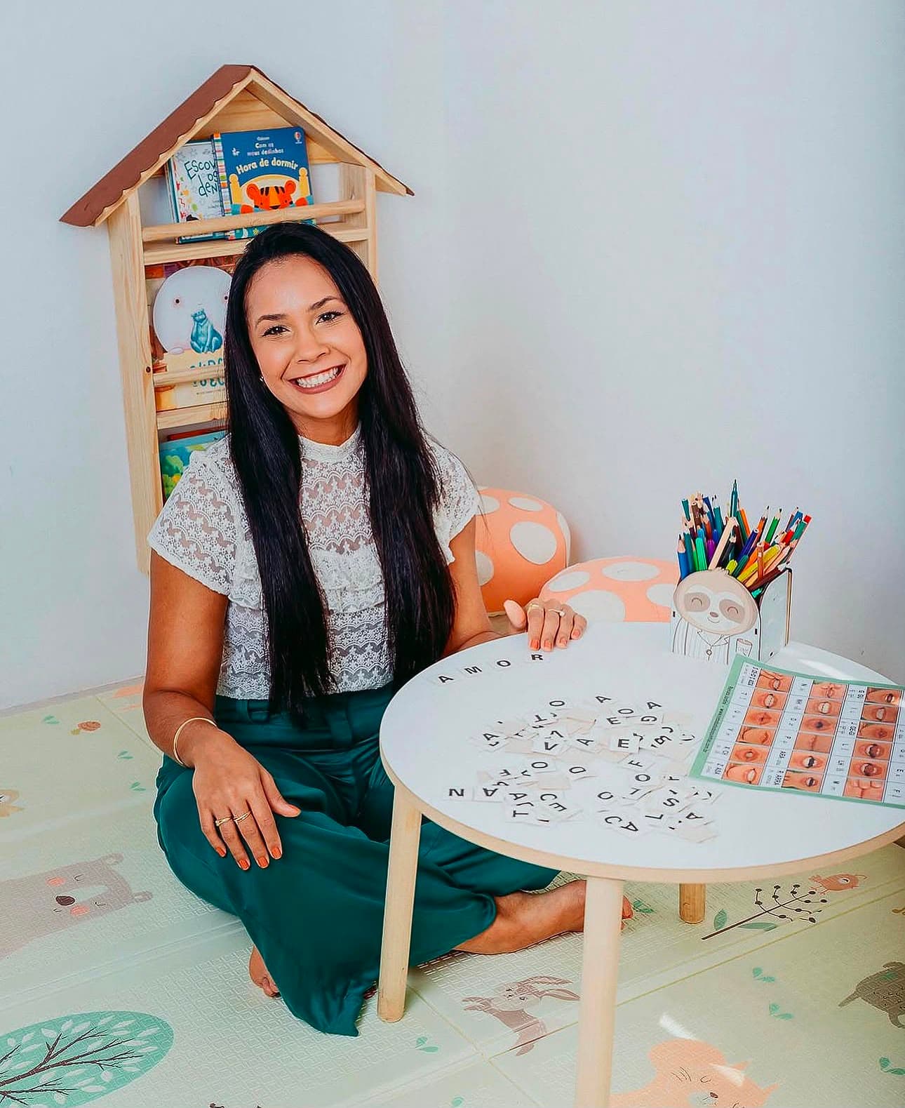 Stella Costa, fonoaudióloga infantil, sorrindo sentada no tapete da sala de atendimento infantil, ao lado de uma mesinha com letras móveis e materiais de terapia de fala